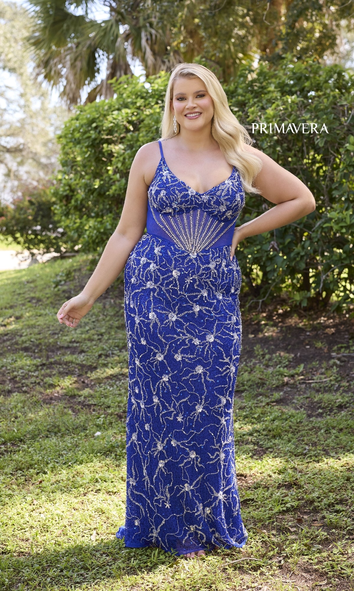 A woman poses outdoors against lush greenery in the Primavera 14079 plus-size long sequin prom gown—a sleeveless, floor-length blue dress with silver starburst sequins on the bodice.